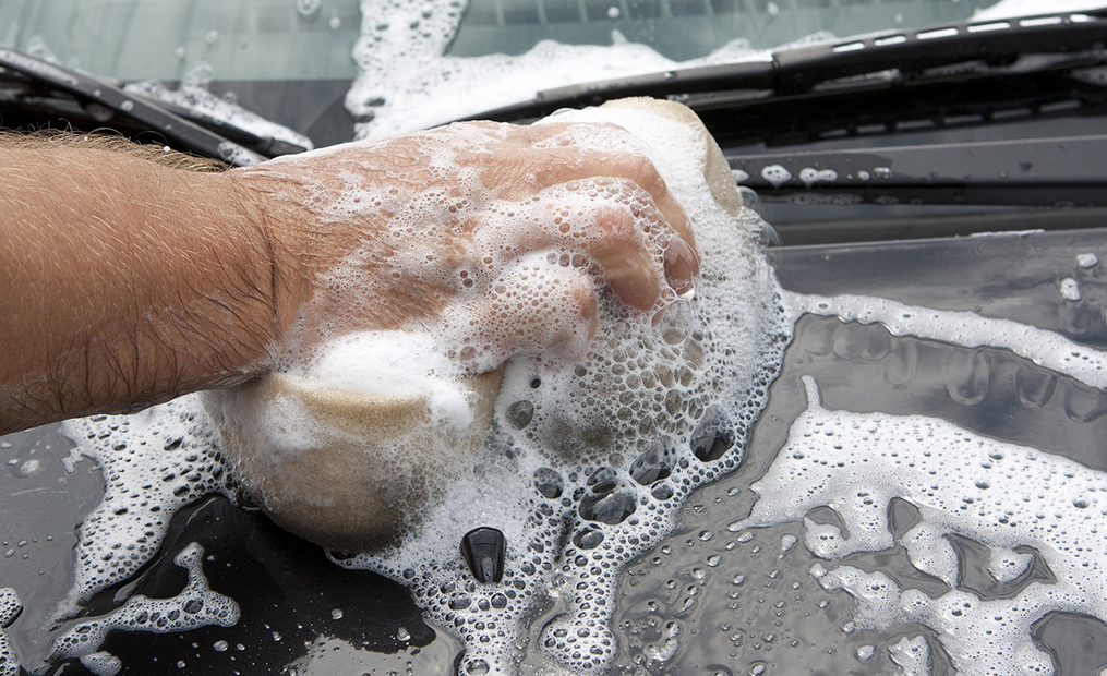 Eine Detailaufnahme einer Männerhand, die mit einem Schwamm gerade die Motorhaube eines Autos einseift