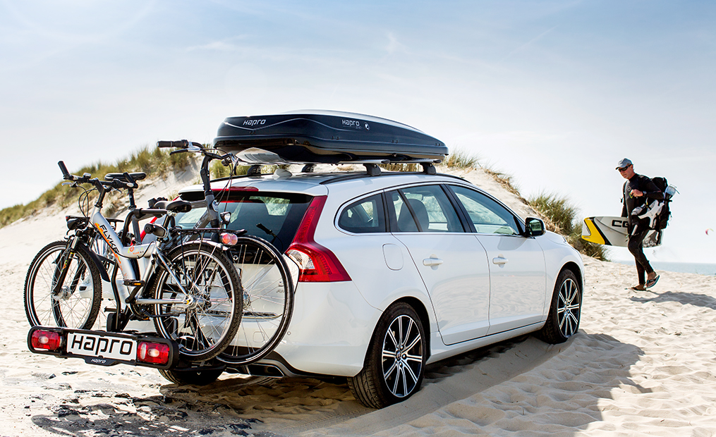 Weißer Kombi am Strand mit zwei Fahrrädern auf einem Anhängerkupplungs-Fahrradträger sowie minimierter Dachbox. Im Hintergrund ein Surfer auf dem Weg zum Auto