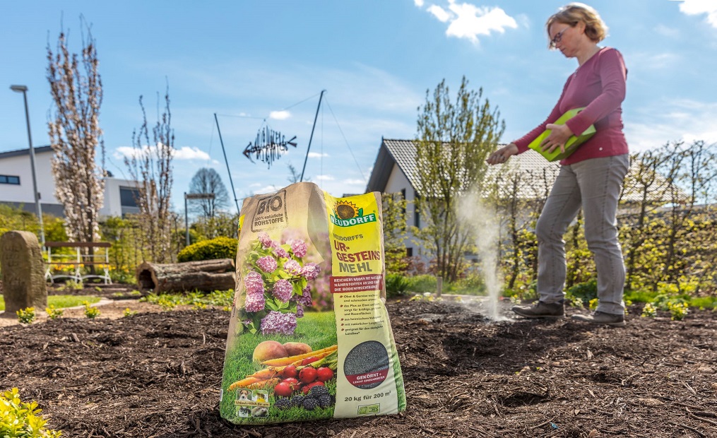 Eine Frau, die Gesteinsmehl auf einem Beet verteilt. Im Hintergrund Häuser und blauer Himmel sowie einige Büsche und eine kleine Hecke
