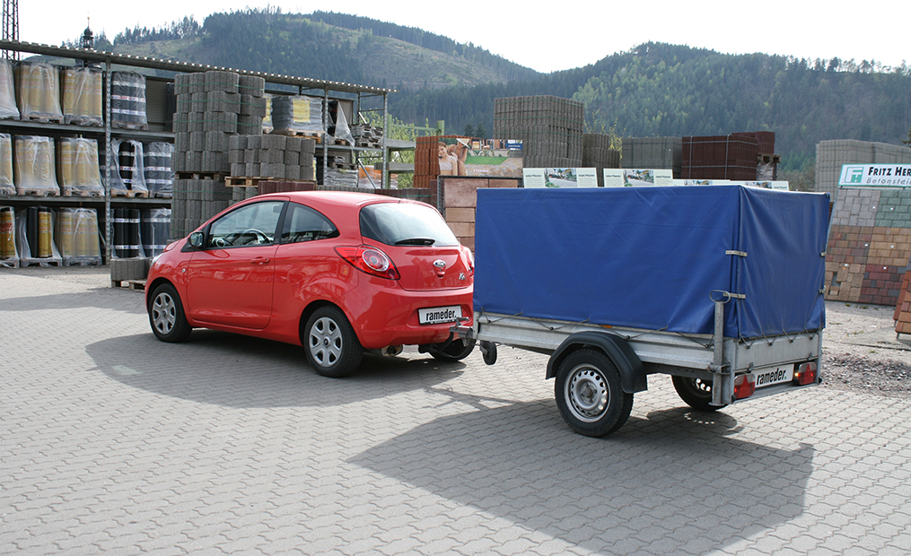 Ein roter Ford Ka mit Anhängerkupplung und kleinem Anhänger. Im Hintergrund Rohre, Betonsteine und weiteres Baumaterial