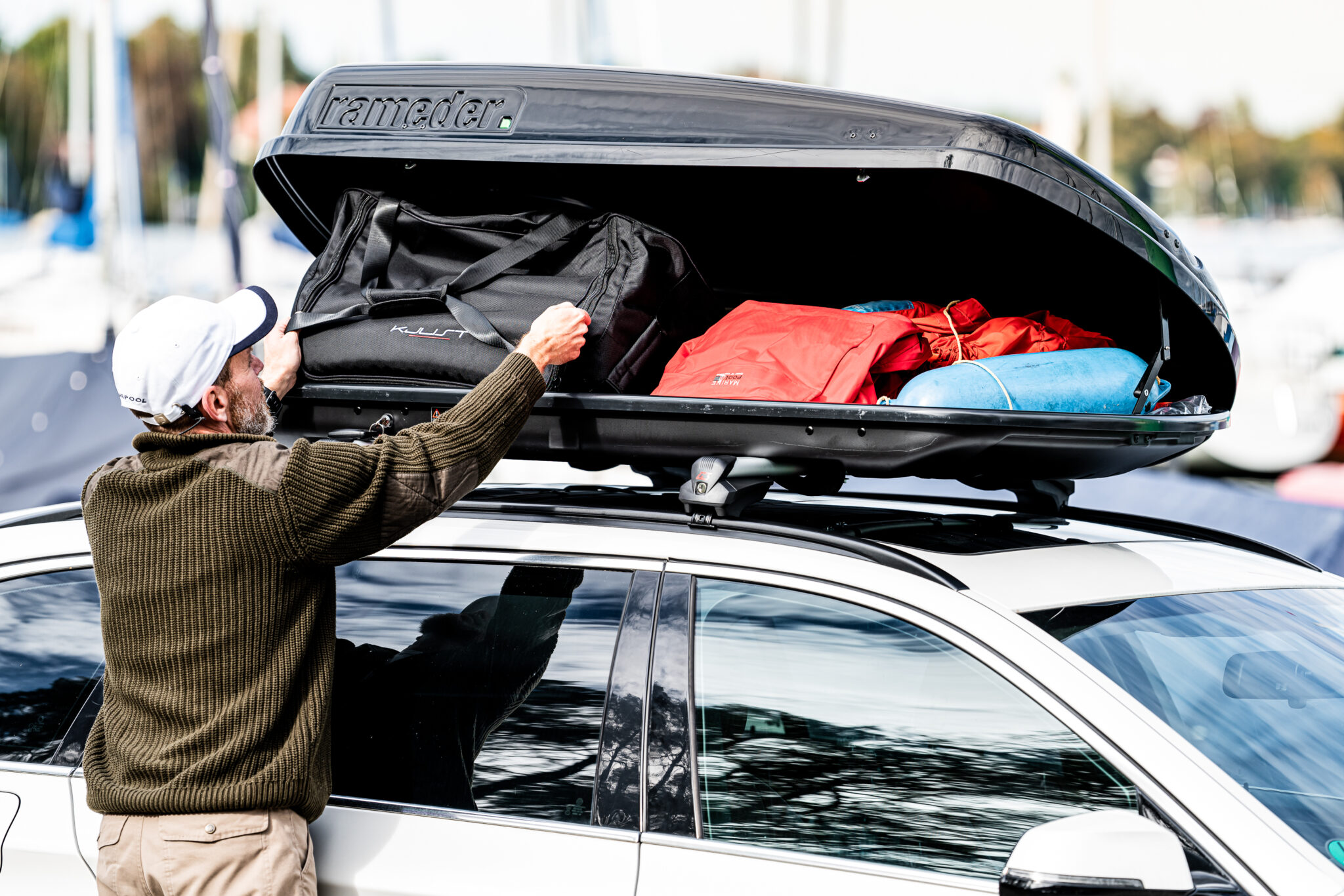 geöffnete Dachbox auf einem Autodach gefüllt mit Taschen ; ein Mann belädt die Box