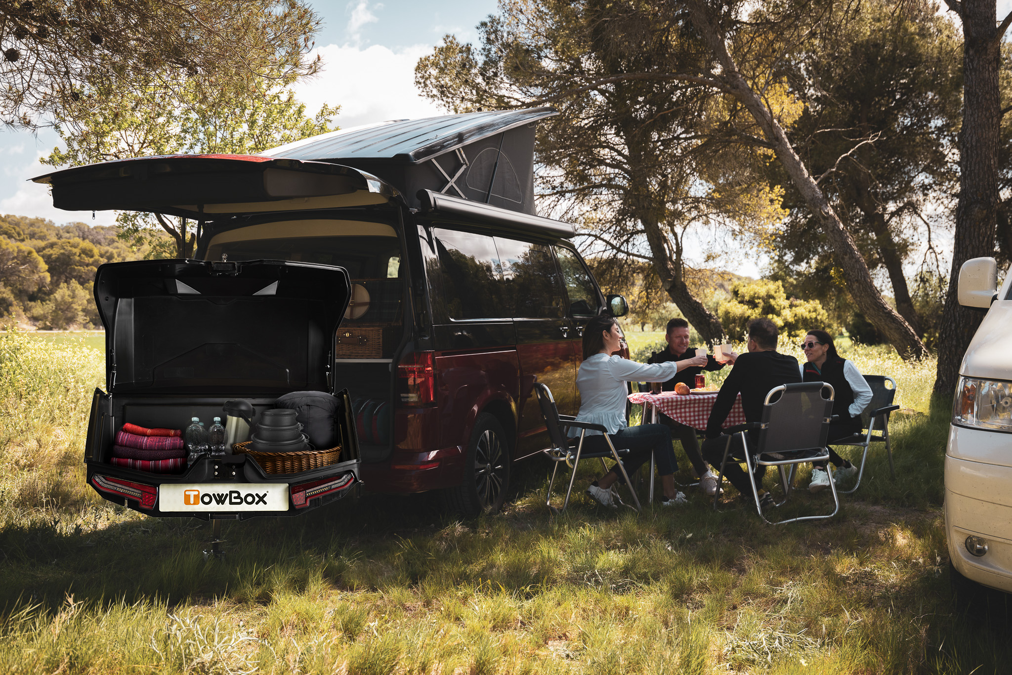 Eine Campingszene: 4 Personen sitzen auf Campingstühlen vor einem Van mit geöffneter Heckklappe, montierter TocwBox V5 Swing sowie montiertem Dachzelt.